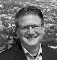 En person i glasögon och kostym ler framför en stadsbakgrund.