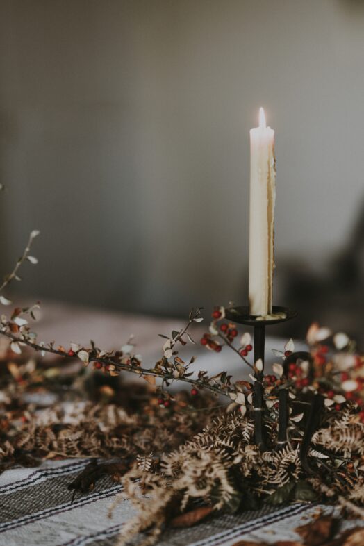 Ett tänt ljus på en ljusstake omgiven av vissna blommor.